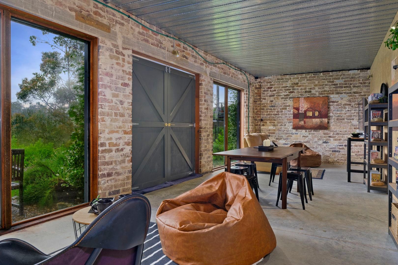 A rustic room crafted with premium reclaimed building materials features brick walls, large windows, a wooden table with chairs, bean bags, and a bookshelf. There's a grand door and a painting adorning one wall.