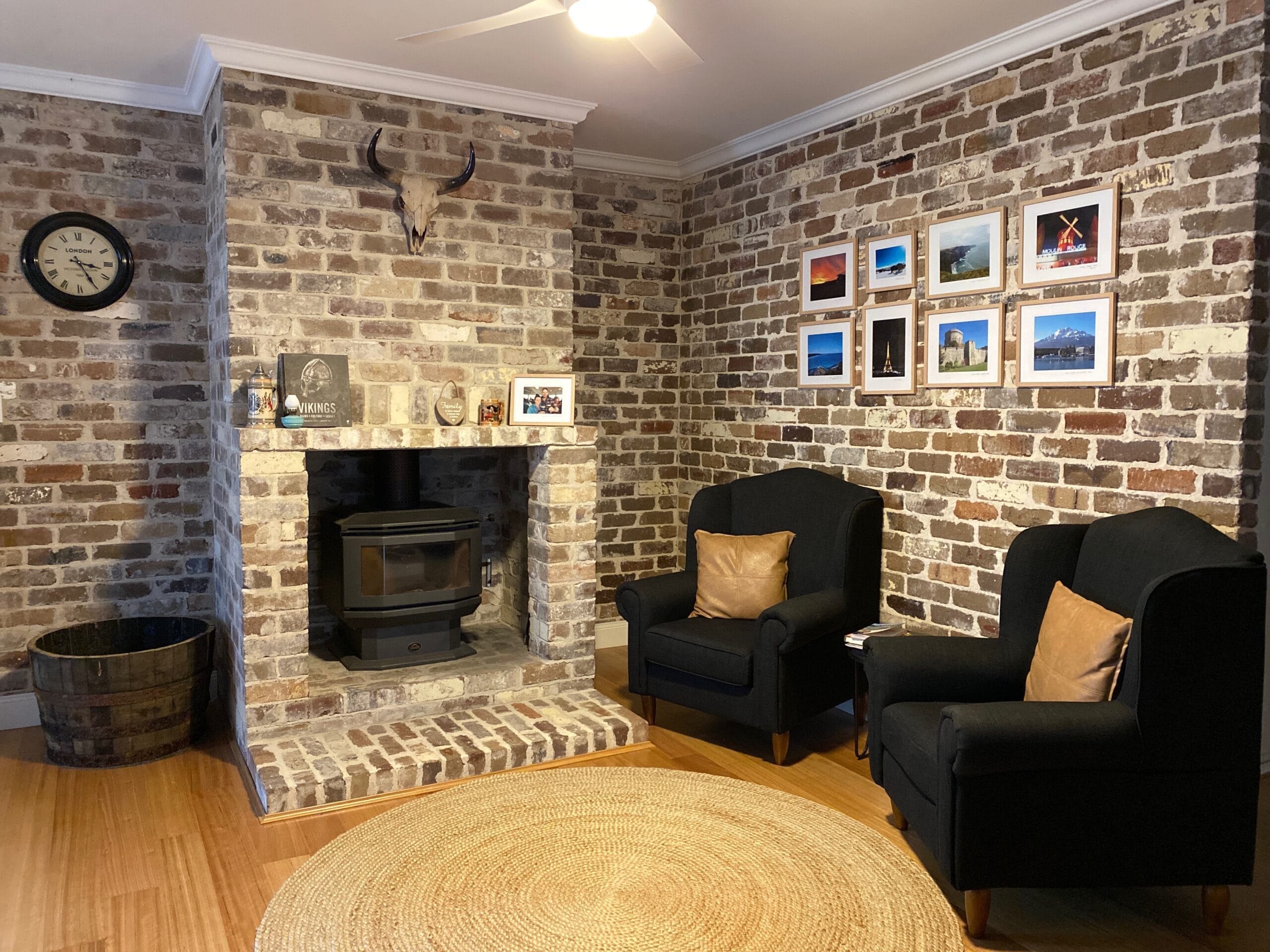 Cozy living room with recycled brick walls, two black armchairs, a wood stove, framed photos, and a decorative animal skull above the fireplace. A round woven rug is on the floor.