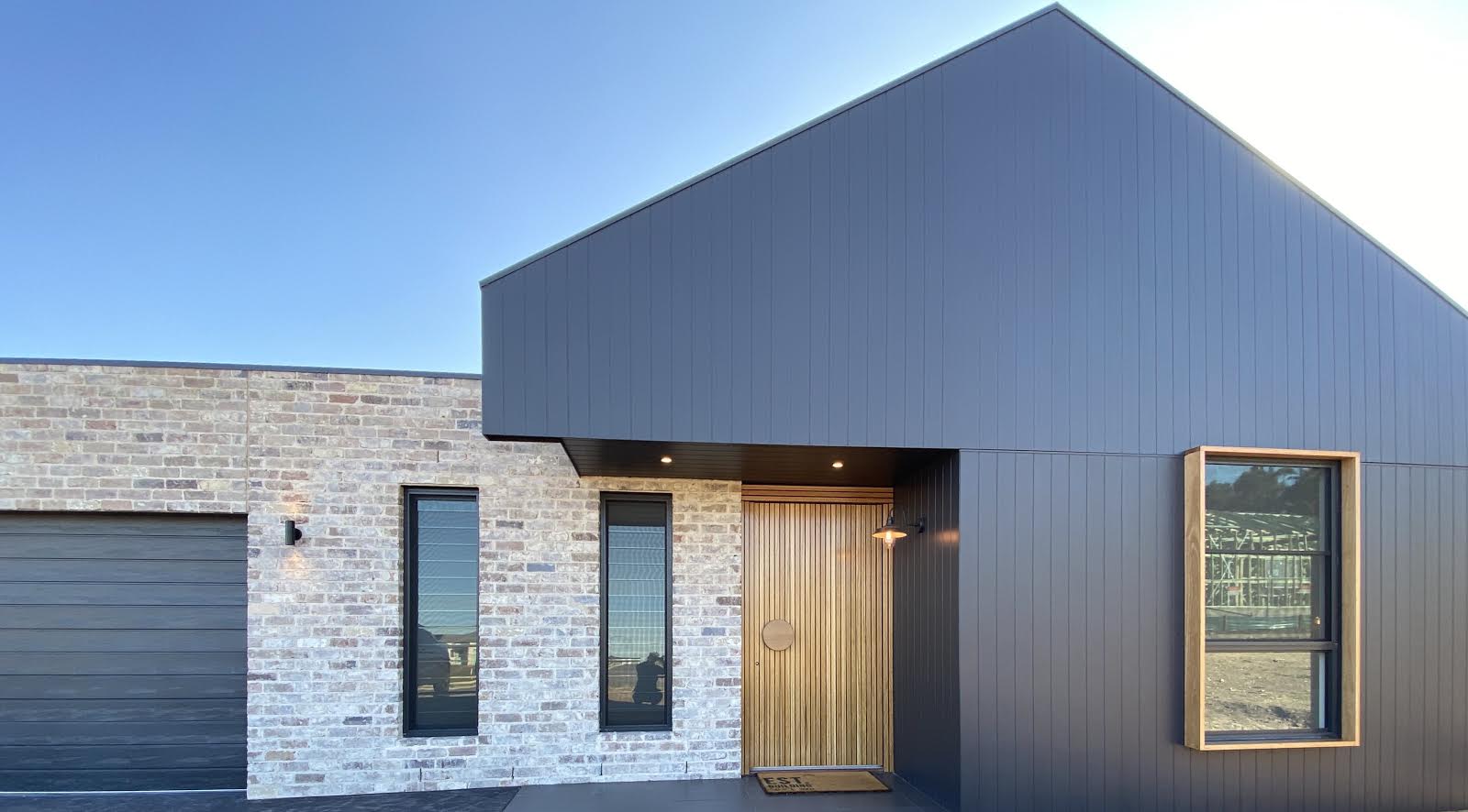 Modern house exterior with a mix of brick and sleek black siding, featuring two tall windows crafted from premium reclaimed materials, a wooden door, and a garage door on the left.