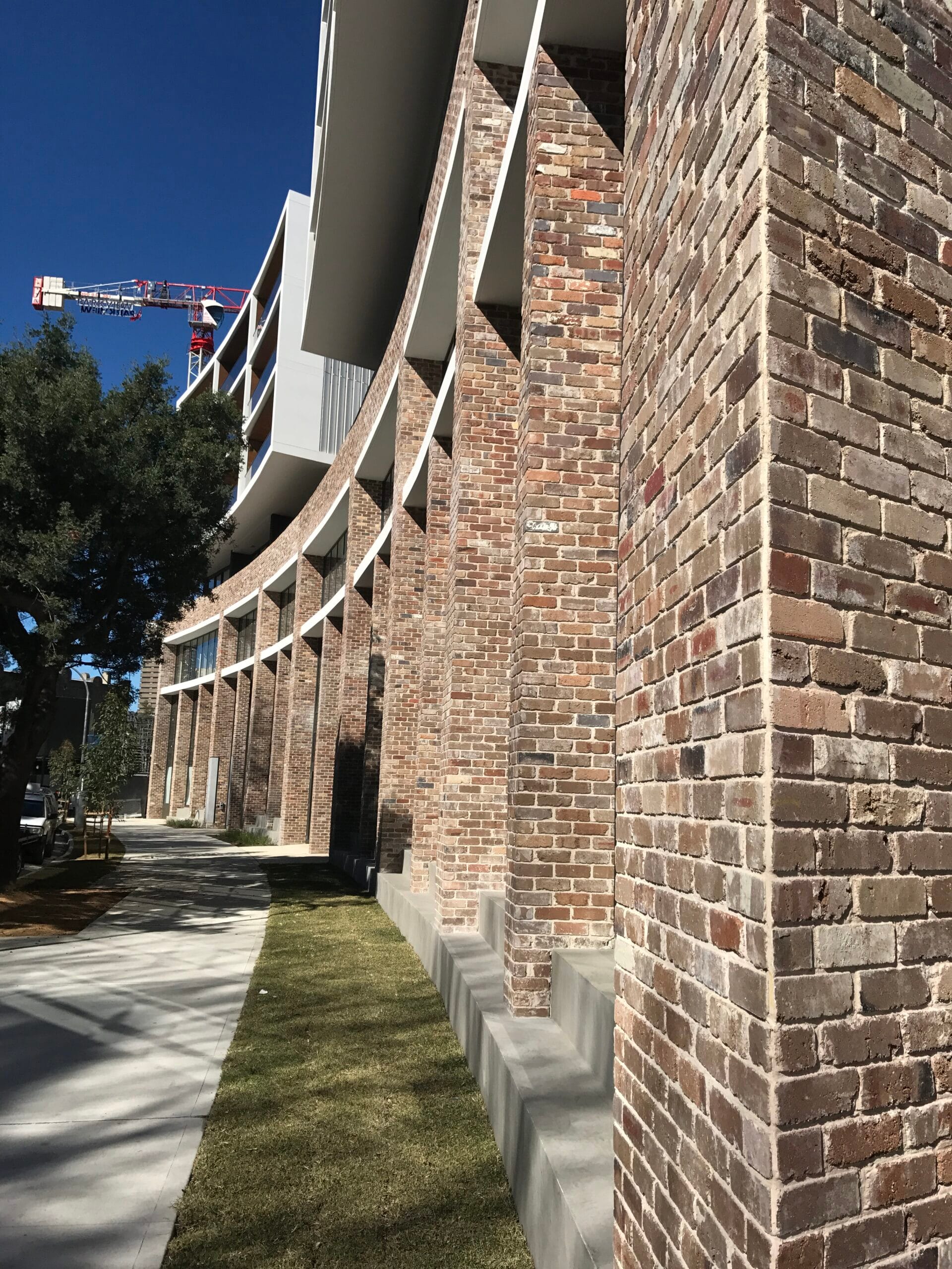 The brick building facade of Parc Mezzo features tall columns and a graceful curved design, complemented by a lush grassy area. In the background, a construction crane stands tall against the clear blue sky.