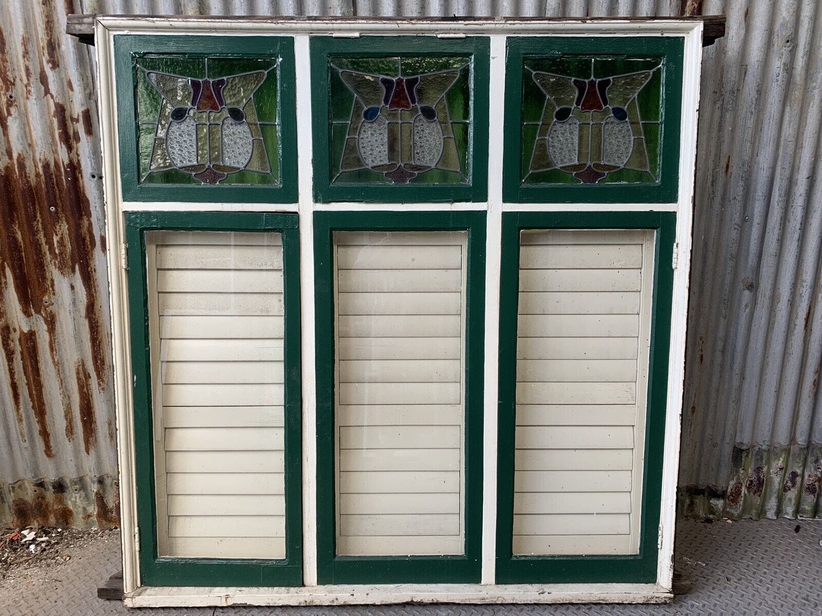 A green and white stained glass window with three panels, featuring geometric designs at the top, crafted from premium reclaimed building materials, leans elegantly against a corrugated metal wall.