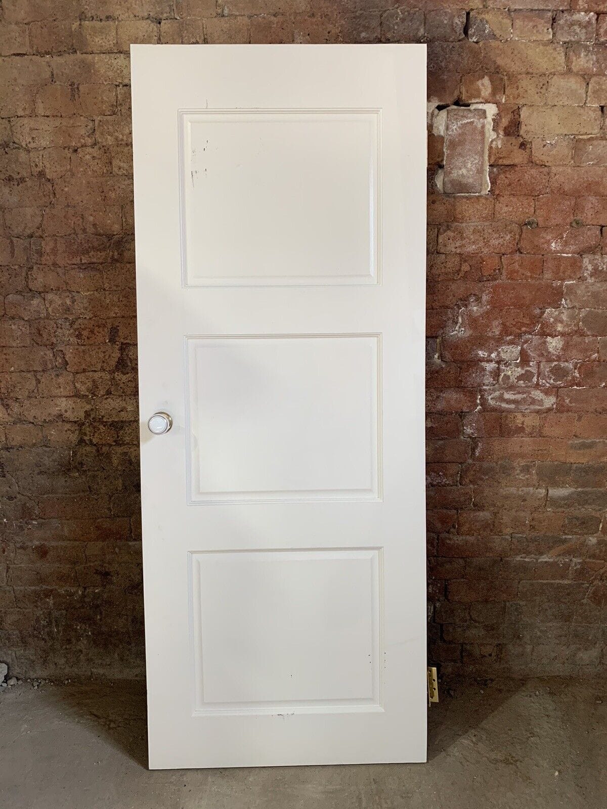 A white three-panel door, crafted from premium reclaimed building materials, stands upright with a round knob against a textured brick wall.