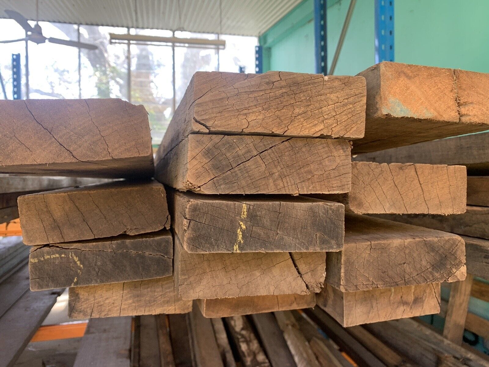 A stack of rough-cut wooden beams, crafted from premium reclaimed building materials, is arranged in a storage area with light streaming in from a window in the background.