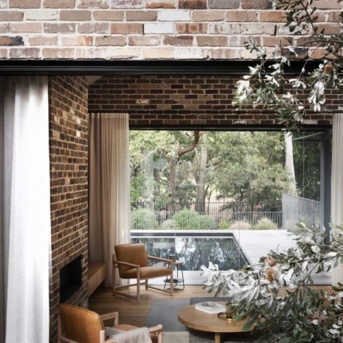 A cozy brick-walled room crafted with premium reclaimed building materials features large windows that open to a stunning view of a pool and lush trees. Two chairs and a table grace the foreground, with elegant curtains drawn back to welcome the serene vista.