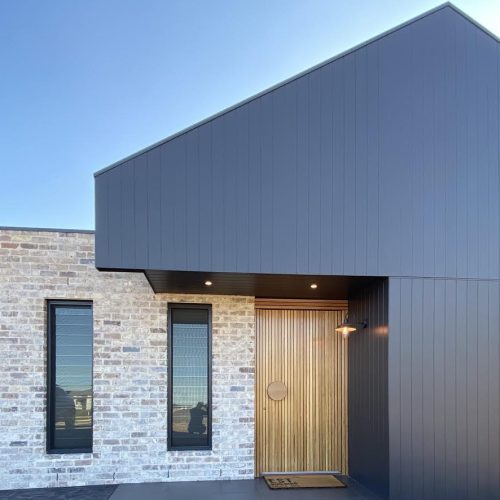 Modern house exterior with a mix of brick and sleek black siding, featuring two tall windows crafted from premium reclaimed materials, a wooden door, and a garage door on the left.