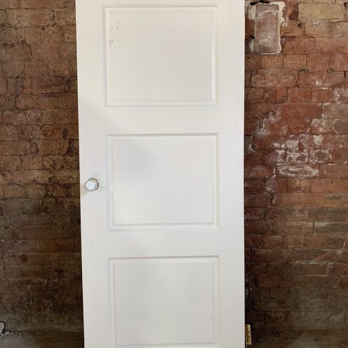 A white three-panel door, crafted from premium reclaimed building materials, stands upright with a round knob against a textured brick wall.