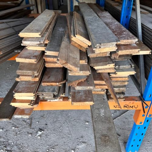 Stack of weathered wooden planks on metal shelves in a storage area. Dust covers the floor and shelves.